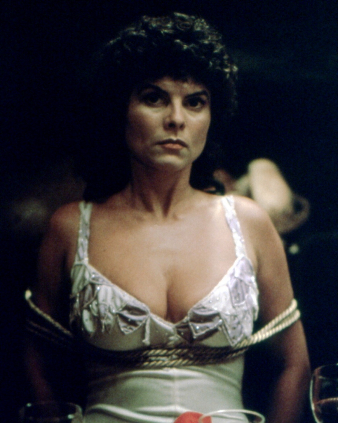 Adrienne Barbeau with an angry face while tied with a rope, and wearing a white sleeveless dress in a movie scene from Swamp Thing, a 1982 American superhero horror film.