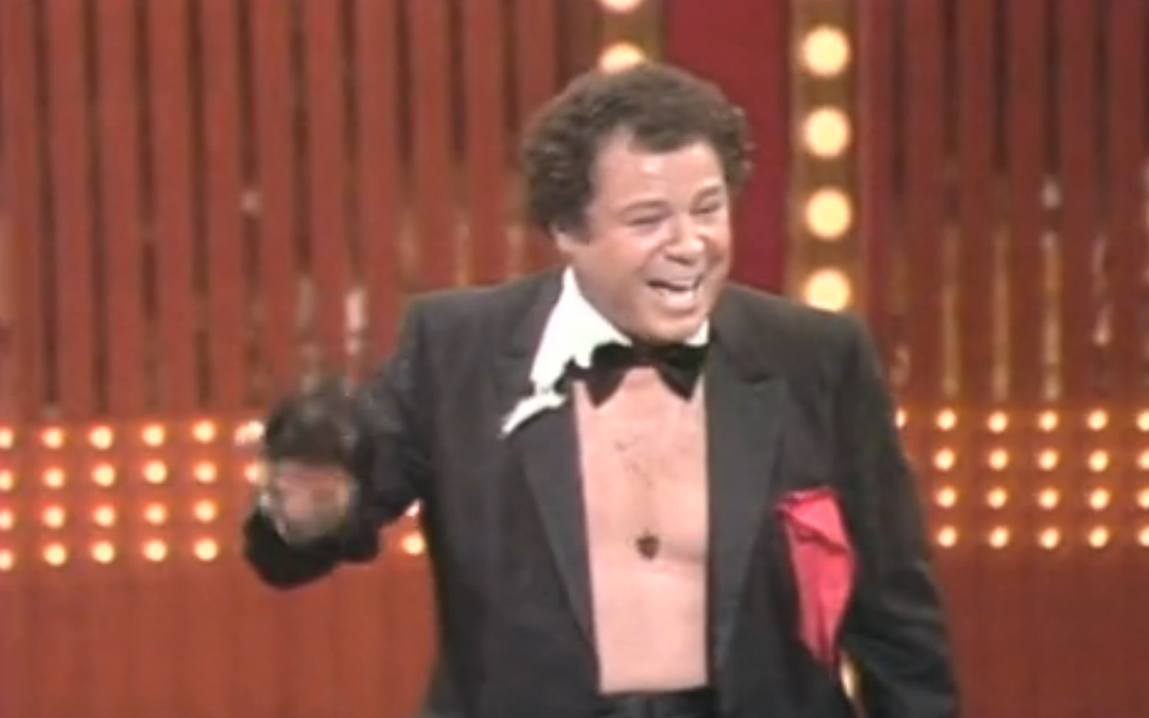 Art Metrano laughing while performing a magic on stage and wearing a black coat and a bow tie