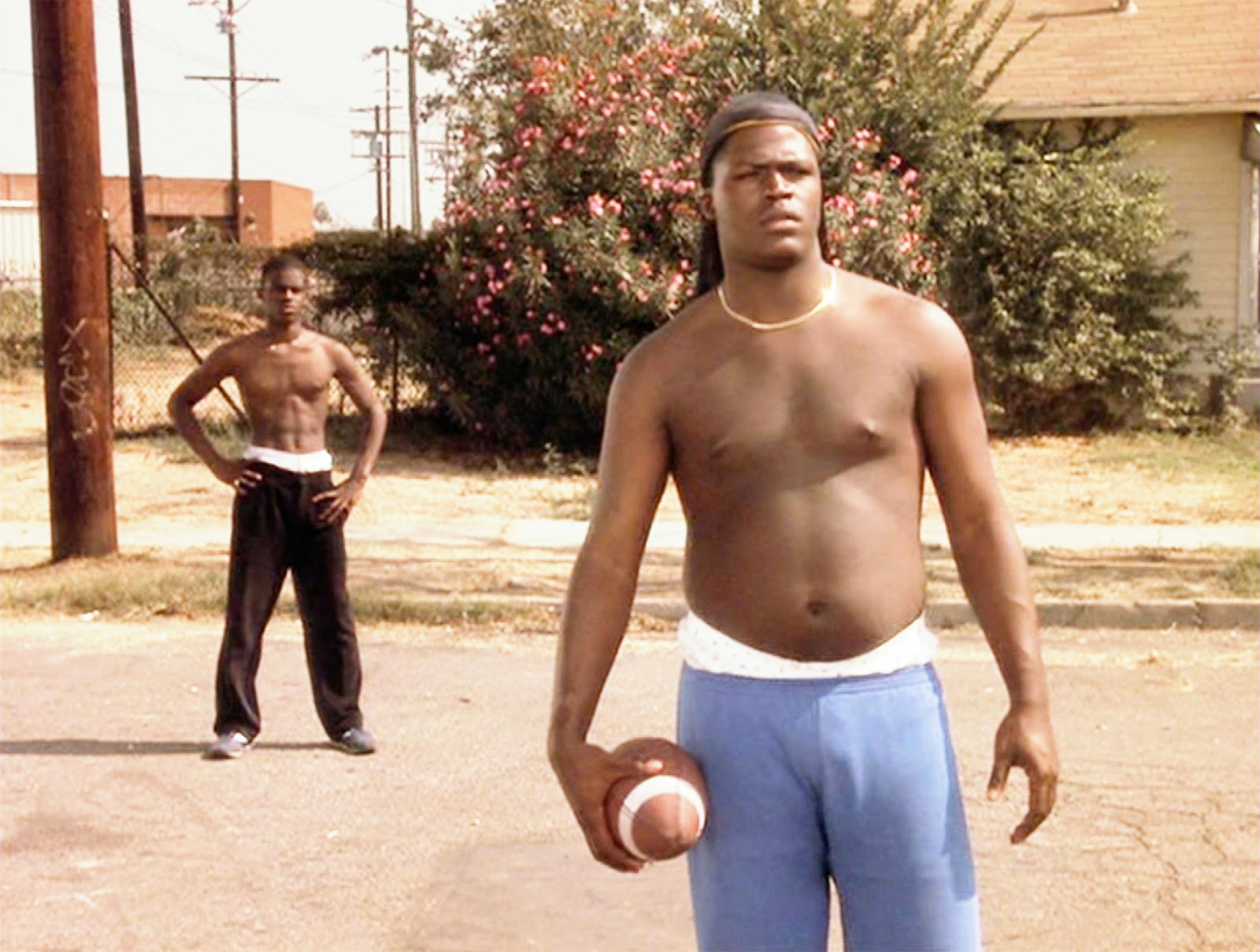 Lexie Bigham wearing brown headdress, gold necklace and blue pants while holding a ball and a man behind him wearing black pants while hands on his hips